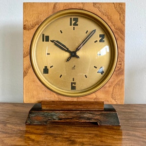 May include: A vintage wooden clock with a square frame and a gold-colored face. The clock features black numerals and hands, with the brand name "JAZ" visible. The clock sits on a rectangular wooden base, showcasing a classic design.
