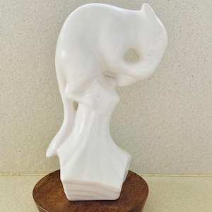 Puede incluir: Figurilla de cerámica blanca de un gato sentado sobre una base de madera. El gato está enroscado con la cabeza apoyada en sus patas.