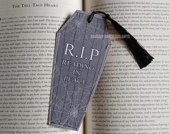 Może przedstawiać: Zakładka do książki w kształcie trumny w odcieniach szarości z białym wzorem pajęczyny i napisem R.I.P READING IN PEACE. Zakładka posiada czarny chwost przymocowany u góry za pomocą małego czarnego sznurka, leżąca na stronie otwartej książki.