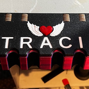 May include: A black and red 3D printed coin holder with the name "TRACI" and a white heart with wings on top. The holder has five slots for coins.