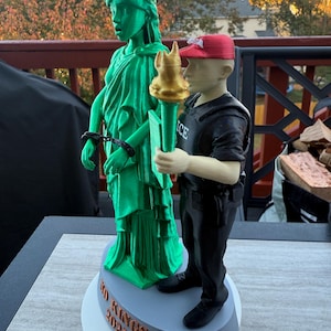 Może przedstawiać: Wydrukowana w 3D rzeźba przedstawiająca Statuę Wolności w kolorze zielonym z kajdankami, obok postaci w czarnym mundurze i czerwonej czapce trzymającej pochodnię. Na podstawie napis "NO KINGS 2025".