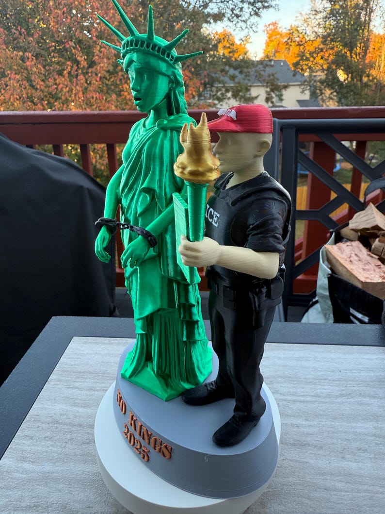 Może przedstawiać: Wydrukowana w 3D rzeźba przedstawiająca Statuę Wolności w kolorze zielonym z kajdankami, obok postaci w czarnym mundurze i czerwonej czapce trzymającej pochodnię. Na podstawie napis "NO KINGS 2025".