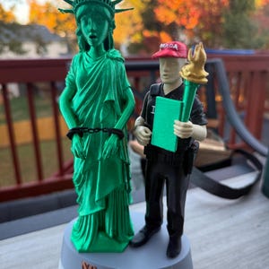 Może przedstawiać: Zielona figurka Statuy Wolności wydrukowana w 3D, z kajdankami i napisem "NO KINGS 2025" na podstawie. Obok stoi postać w czapce "MAGA" trzymająca pochodnię i zielony notatnik.