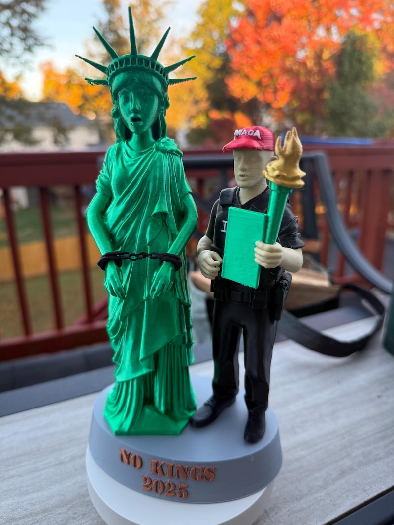 Może przedstawiać: Zielona figurka Statuy Wolności wydrukowana w 3D, z kajdankami i napisem "NO KINGS 2025" na podstawie. Obok stoi postać w czapce "MAGA" trzymająca pochodnię i zielony notatnik.