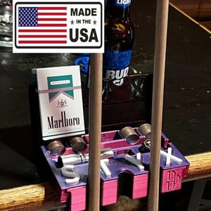 May include: A purple and pink 3D printed pool cue rack with the word "STYX" and the letters "DB" on the side. The rack has slots for pool cues, a cigarette pack, and a bottle. The rack is sitting on a wooden table with a Bud Light beer bottle and a pack of Marlboro cigarettes.