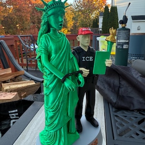 Może przedstawiać: Zielona figurka Statuy Wolności z kajdankami, obok figurki osoby w czerwonej czapce "MAGA" i kamizelce "ICE", trzymającej zielony tablet. Na podstawie napis "NO RINGS 2025".