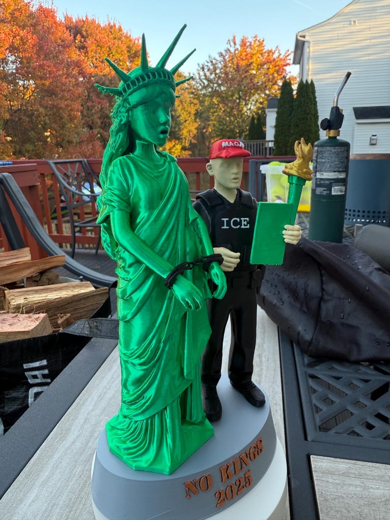 Może przedstawiać: Zielona figurka Statuy Wolności z kajdankami, obok figurki osoby w czerwonej czapce "MAGA" i kamizelce "ICE", trzymającej zielony tablet. Na podstawie napis "NO RINGS 2025".