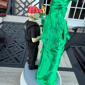 Może przedstawiać: 3D-wydrukowana figurka Statuy Wolności, zielona ze złotą pochodnią, oraz postać w czerwonej czapce i czarnym mundurze. Figurka jest zamontowana na wielowarstwowej podstawie. Statua jest symbolem wolności.