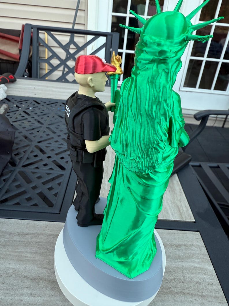 Może przedstawiać: 3D-wydrukowana figurka Statuy Wolności, zielona ze złotą pochodnią, oraz postać w czerwonej czapce i czarnym mundurze. Figurka jest zamontowana na wielowarstwowej podstawie. Statua jest symbolem wolności.