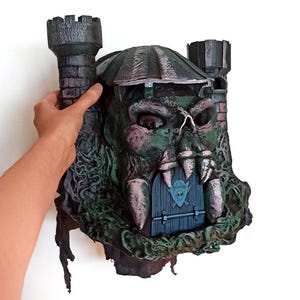 May include: A detailed, hand-painted sculpture of a fantasy castle, featuring a skull-like facade with large fangs and a blue door. The castle has two turrets and is primarily green, with accents of pink and black.