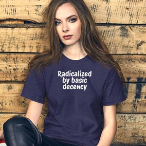 Könnte beinhalten: Ein marineblaues T-Shirt mit dem weißen Text "Radicalized by basic decency". Das kurzärmelige Shirt hat einen Rundhalsausschnitt. Der Hintergrund ist eine Holzwand.