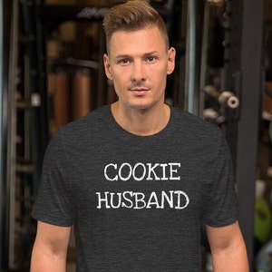 Könnte beinhalten: Ein dunkelgraues T-Shirt mit dem Aufdruck "COOKIE HUSBAND" in Weiß.