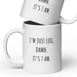 May include: Two white ceramic mugs stacked together. The bottom mug has the text "I'M JUST LIKE, DAMN. IT'S 7 AM." in black. The top mug has the same text. Both mugs have a curved handle.