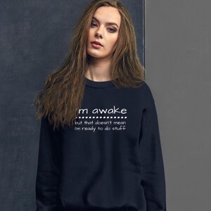 May include: A navy blue sweatshirt with white text reading "I'm awake but that doesn't mean I'm ready to do stuff". The crew neck sweatshirt is made of a soft material.