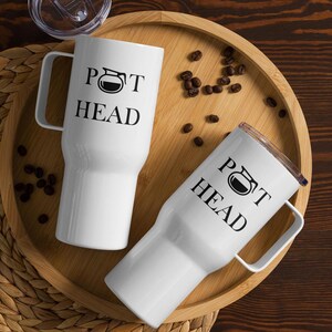 Könnte beinhalten: Zwei weiße Reisebecher mit schwarzen Griffen und dem Text "POT HEAD" und einer schwarzen Grafik einer Kaffeekanne. Die Becher befinden sich auf einem Holztablett mit verstreuten Kaffeebohnen.