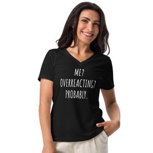 May include: Black V-neck t-shirt with the text "ME? OVERREACTING? PROBABLY." in white. The shirt is made of a soft material and has short sleeves. The person is smiling.