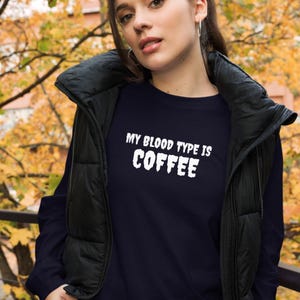 May include: A navy blue long-sleeve shirt with the white text "MY BLOOD TYPE IS COFFEE". The shirt is worn with a black vest. The background features autumnal foliage.