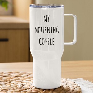 Könnte beinhalten: Ein weißer Reisebecher mit Henkel und transparentem Deckel. Der Becher trägt den schwarzen Aufdruck "MY MOURNING COFFEE". Der Becher ist zylindrisch mit leicht konischem Boden. Ideal für heiße oder kalte Getränke.