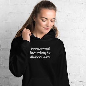 May include: A black hooded sweatshirt with the text "introverted but willing to discuss cats" in white.