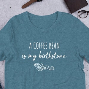 May include: A teal blue t-shirt with the text "A coffee bean is my birthstone" in white lettering. A small graphic of coffee beans is below the text.
