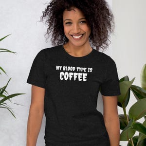 May include: A dark gray t-shirt with the text "MY BLOOD TYPE IS COFFEE" in white block letters. The shirt is being worn by a person with curly dark hair.