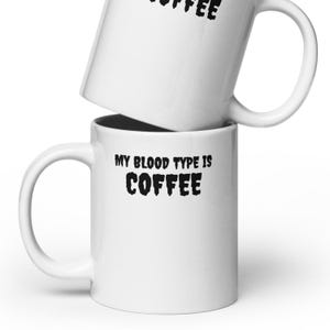 May include: Two white ceramic coffee mugs stacked on top of each other. The bottom mug has the text "MY BLOOD TYPE IS COFFEE" in black, bold letters. The top mug has the word "COFFEE" in black, bold letters.