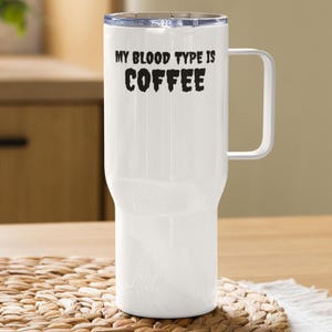 Peut inclure: Mug de voyage blanc avec une poignée et un couvercle transparent. Le mug porte l'inscription "MY BLOOD TYPE IS COFFEE" en lettres noires. Le mug est posé sur un dessous de plat tissé.