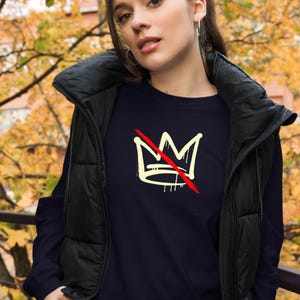 May include: Navy blue long-sleeve shirt featuring a cream-colored crown design with a red slash through it. The graphic tee is a casual style, suitable for everyday wear. The shirt is paired with a black vest.