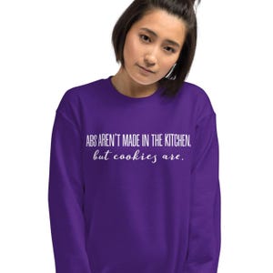 May include: A purple sweatshirt with the text "ABS AREN'T MADE IN THE KITCHEN, but cookies are." The text is in a white, handwritten-style font. The sweatshirt is a solid color and appears to be made of a soft material.