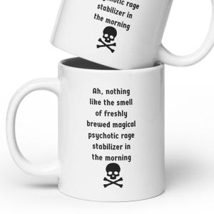 May include: White ceramic mug with black text and a skull and crossbones design. The text reads "Ah, nothing like the smell of freshly brewed magical psychotic rage stabilizer in the morning."