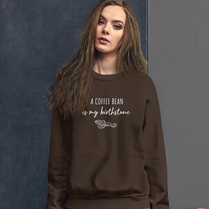 以下が含まれることがあります： 「A coffee bean is my birthstone」の白い文字がプリントされた茶色のスウェットシャツ。 下には3つのコーヒー豆のイラストがあります。 このスウェットシャツは無地で、クルーネックです。