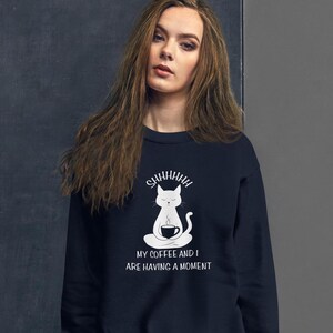 May include: A navy blue sweatshirt with a white graphic of a cat meditating with a cup of coffee. The text on the sweatshirt reads "Shhhhh My coffee and I are having a moment."