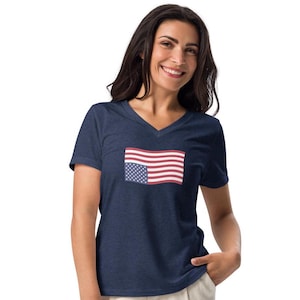 May include: A navy blue V-neck t-shirt featuring a small American flag graphic on the front. The flag design includes red and white stripes and a blue field with white stars. The shirt is made of a soft, comfortable material.