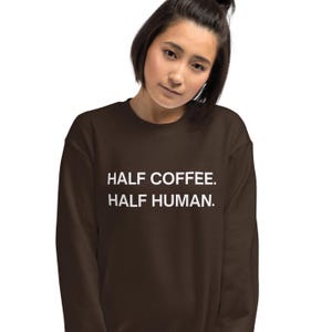 May include: A dark brown sweatshirt with the text "HALF COFFEE. HALF HUMAN." in white. The sweatshirt is a solid colour and has a crew neck. The image shows a person wearing the sweatshirt.