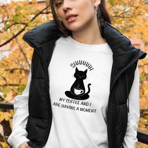 May include: White long-sleeved shirt featuring a black cat illustration holding a cup of coffee. The text "Shhhhhhh My coffee and I are having a moment" is printed on the shirt.
