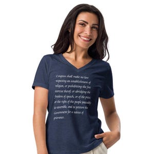 May include: A navy blue v-neck t-shirt with a white print of the First Amendment to the United States Constitution. The text reads: "Congress shall make no law respecting an establishment of religion, or prohibiting the free exercise thereof; or abridging the freedom of speech, or of the press; or the right of the people peaceably to assemble, and to petition the Government for a redress of grievance."