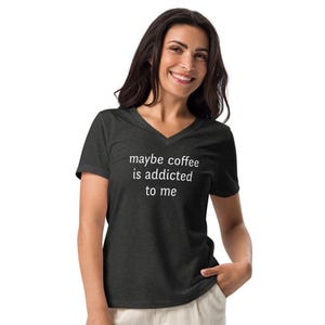 Könnte beinhalten: Dunkelgraues V-Ausschnitt-T-Shirt mit dem weißen Text "maybe coffee is addicted to me". Das Shirt besteht aus einem weichen Material und eignet sich für Freizeitkleidung. Das Model lächelt.