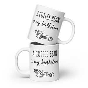 Peut inclure: Deux tasses à café blanches empilées l'une sur l'autre. Les deux tasses ont un texte noir qui dit "A coffee bean is my birthstone" et un graphique de grains de café.