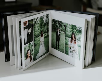 May include: A white wedding album with photos of a bride and groom in a green outdoor setting. The album is open to a page with a collage of photos, including a close-up of the groom in a suit, the bride in a white dress, and a couple standing together.