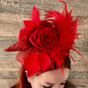 May include: A striking red fascinator with a large rose, feathers, and glitter details. The headpiece features a cluster of red roses and a fan of feathers, suitable for a special event or occasion.