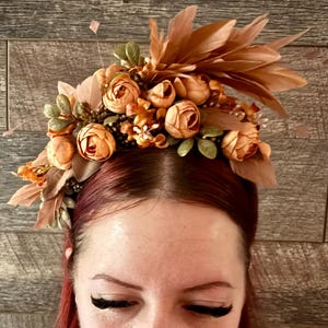 May include: A decorative floral headpiece with peach roses, green leaves, and brown feathers. The headpiece is set against a wooden background. This crown is suitable for weddings, festivals, or special occasions.