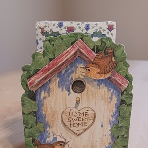 May include: A wooden birdhouse with a red roof and blue siding. The birdhouse has a heart-shaped sign that says "Home Sweet Home". There are two brown birds on the birdhouse, one on each side. The birdhouse is surrounded by green ivy.