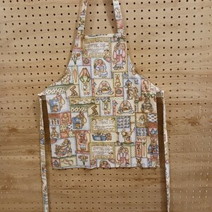 May include: A beige kitchen apron with a patchwork design featuring bunnies, flowers, and vintage-style recipes. The apron has long ties and a hanging loop.