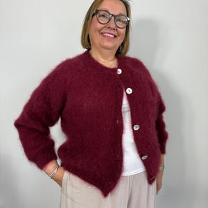 May include: A burgundy cardigan with large white buttons. The fluffy knitwear has long sleeves and is paired with light-colored pants. The cardigan is worn over a white top.