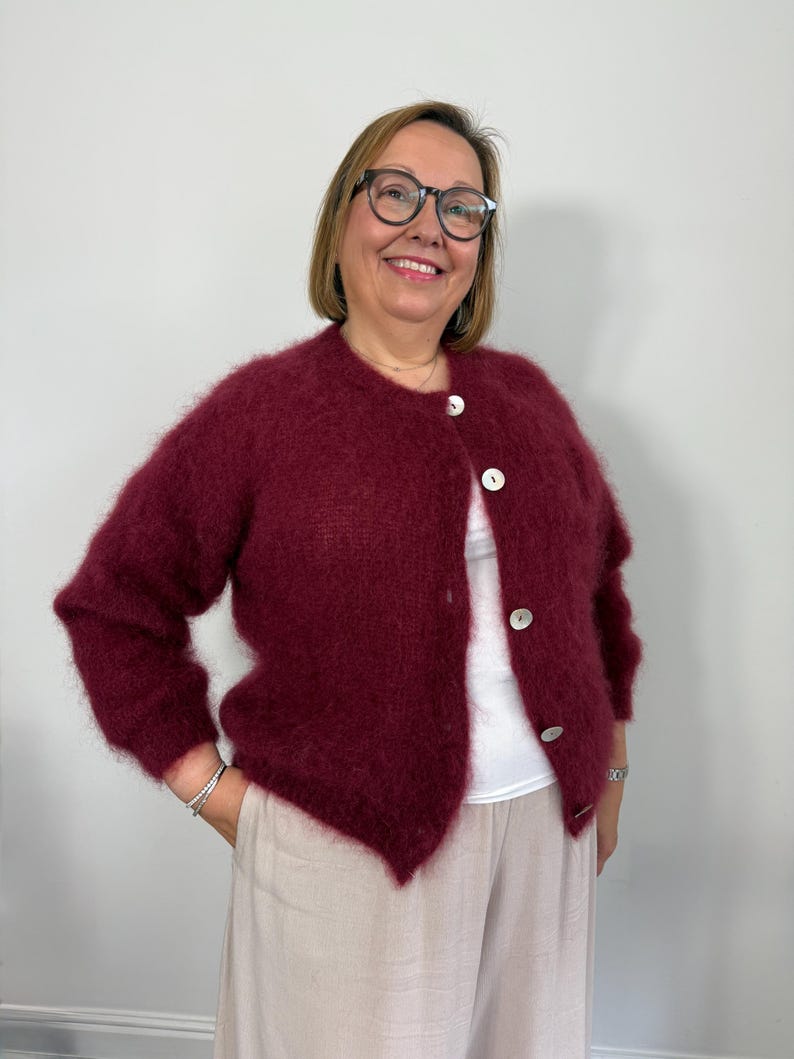 May include: A burgundy cardigan with large white buttons. The fluffy knitwear has long sleeves and is paired with light-colored pants. The cardigan is worn over a white top.