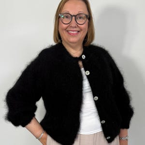 May include: A black, fluffy cardigan with large, round, white buttons. The cardigan has three-quarter sleeves and is worn over a white top and light pink pants. The person in the image is wearing glasses and a watch.