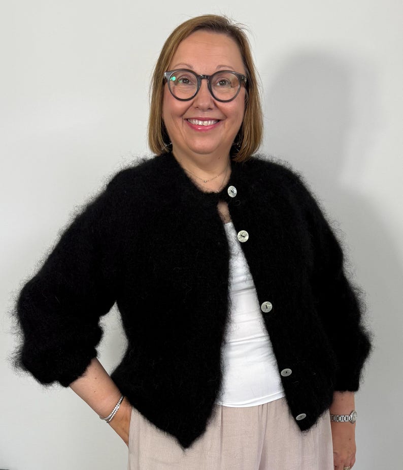 May include: A black, fluffy cardigan with large, round, white buttons. The cardigan has three-quarter sleeves and is worn over a white top and light pink pants. The person in the image is wearing glasses and a watch.