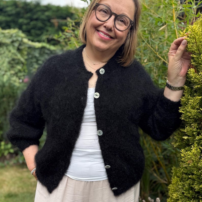 May include: A black, fluffy cardigan with a round neckline and large, pearlescent buttons. The cardigan is worn over a white top and paired with light-colored pants. The image is taken outdoors with greenery in the background.