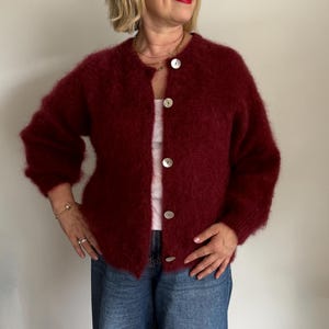 May include: A burgundy, fuzzy cardigan with large, round, white buttons. The cardigan is paired with a white top and blue denim jeans. The woman is wearing a gold necklace and bracelet.