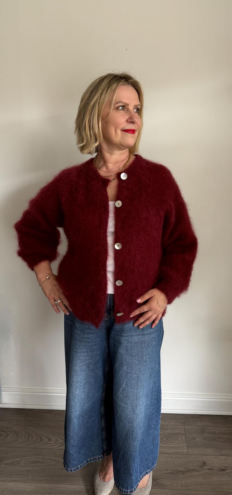 May include: A burgundy, fuzzy cardigan with large, round, white buttons. The cardigan is paired with a white top and blue denim jeans. The woman is wearing a gold necklace and bracelet.